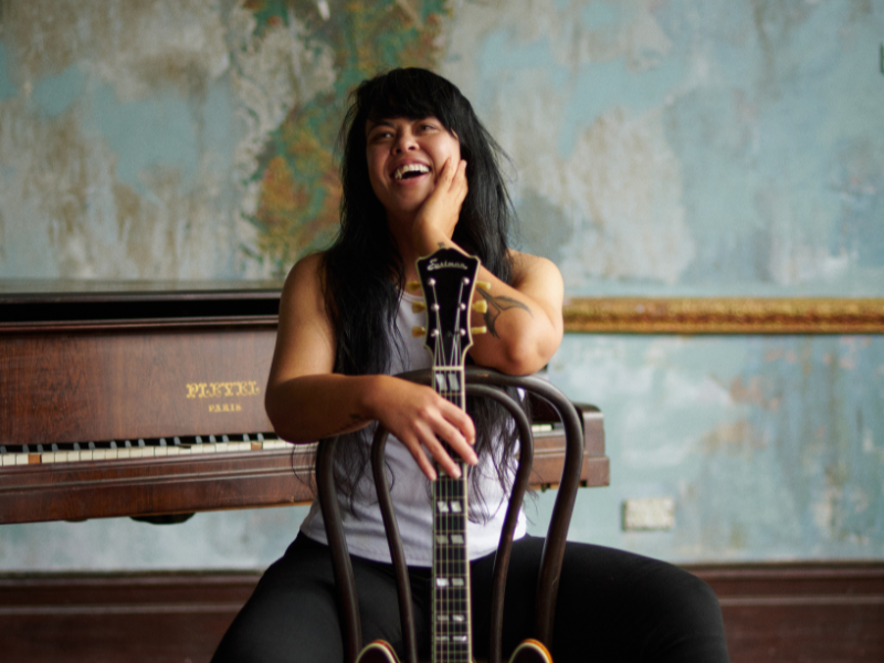 A women sitting backwards on a chair holding an electric guitar and smiling