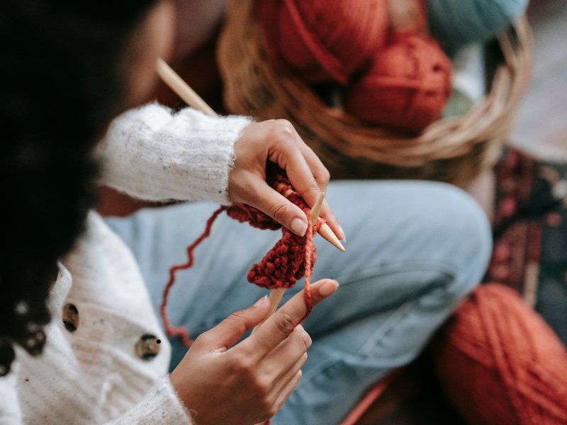 image show an over the shoulder of a woman knitting with red yarn